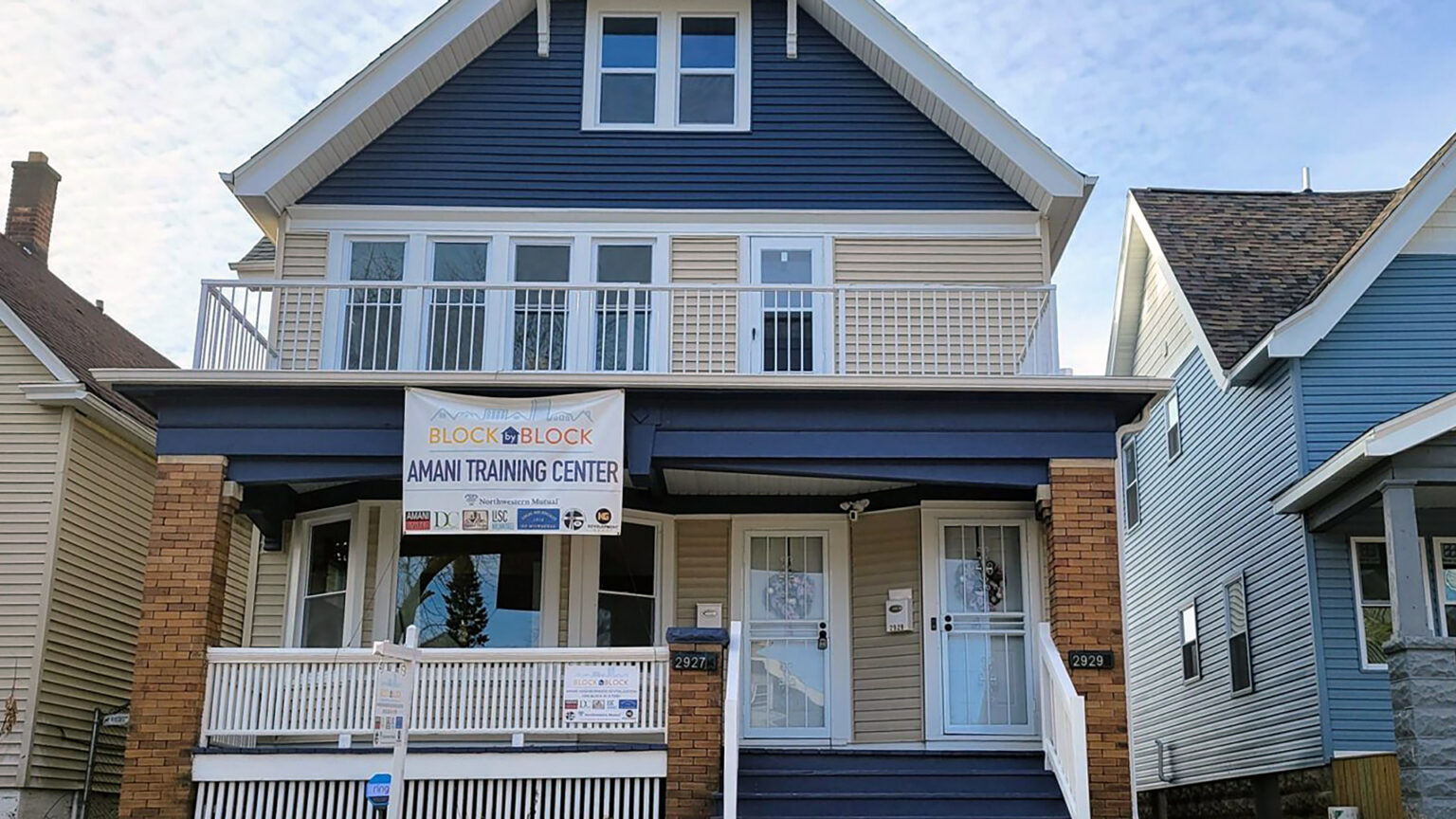 A two-story house with two apartment flats has a banner on its second-floor balcony reading Block by Block Amani Training Center with logos of sponsoring organizations.