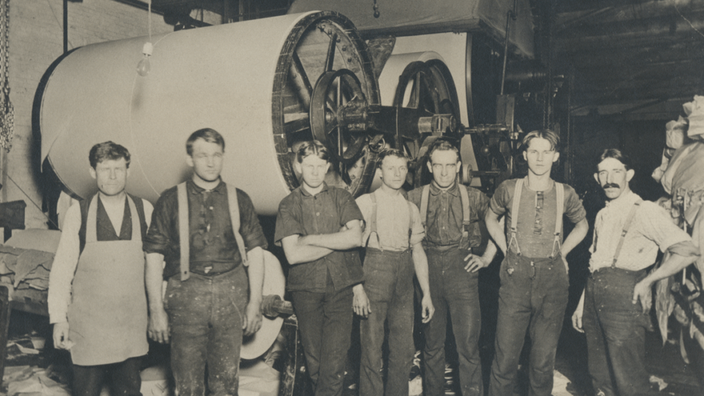 20th century paper mill workers pose in front of large equipment.