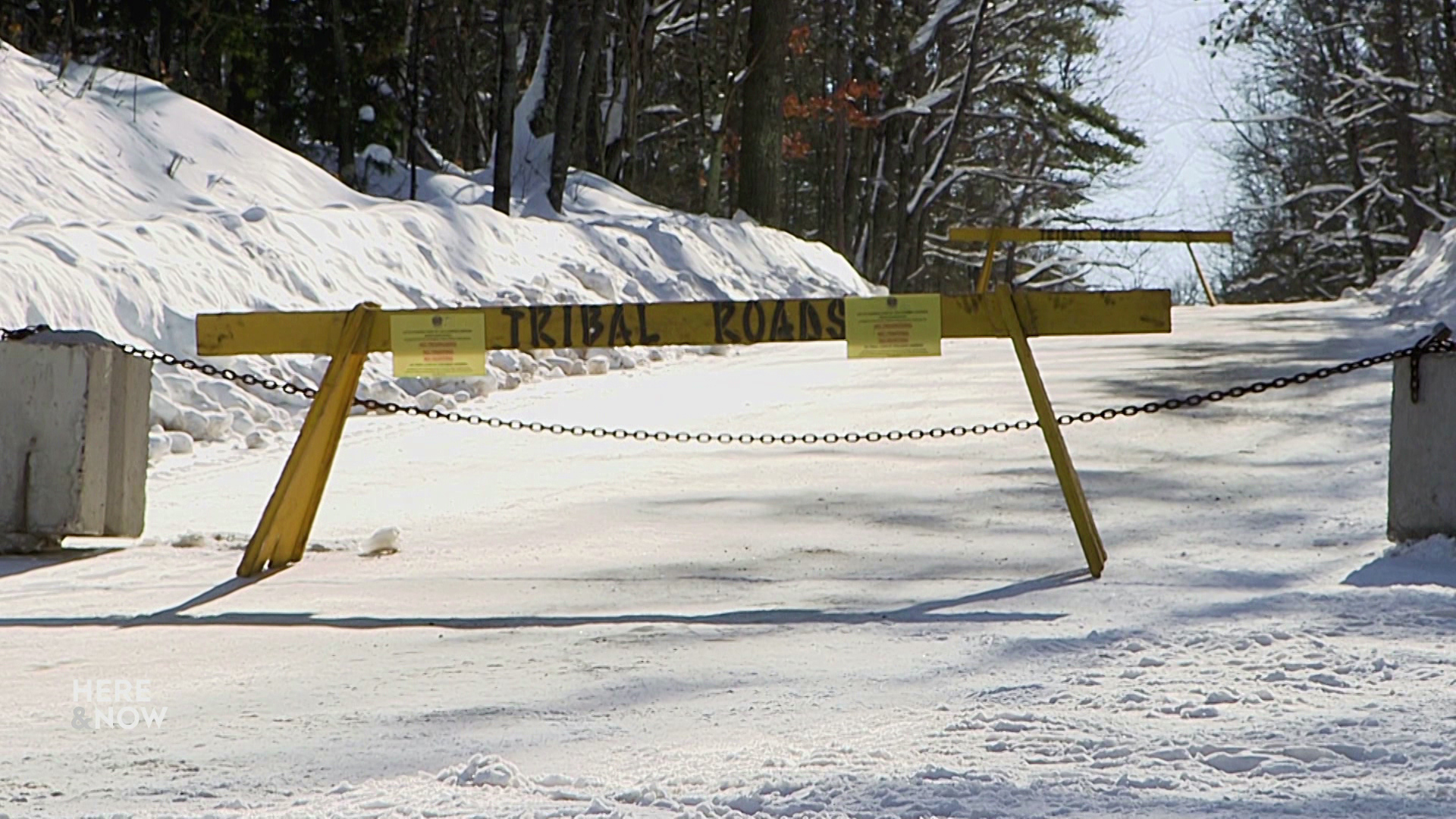 A wood road barrier with "tribal roads" painted on it and two concrete blocks connected with a metal chain sit in front of a road covered in snow, with trees in the background.