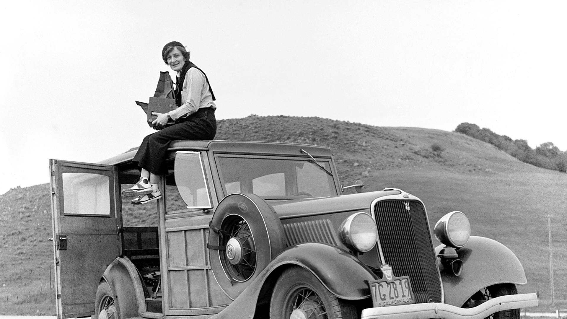 Black and white image of photographer Dorothea Lange sitting on top of a truck on a California highway. She is looking at a map.