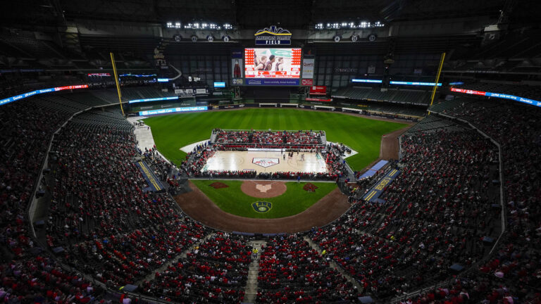 A temporary basketball court is set up on the infield of a baseball stadium, with fans in the first two levels of seating and the game displayed on a large screen situated above center field.