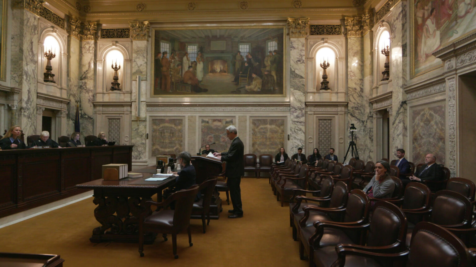 A man stands behind a podium and looks at papers while facing a judicial bench with multiple seated justices, with other people seated at tables on either side of the podium and in rows of leather-and-wood chairs facing the dais and on a side wall, in an ornate room with marble masonry, electrified lighting in wall niches, crown moulding and paintings.