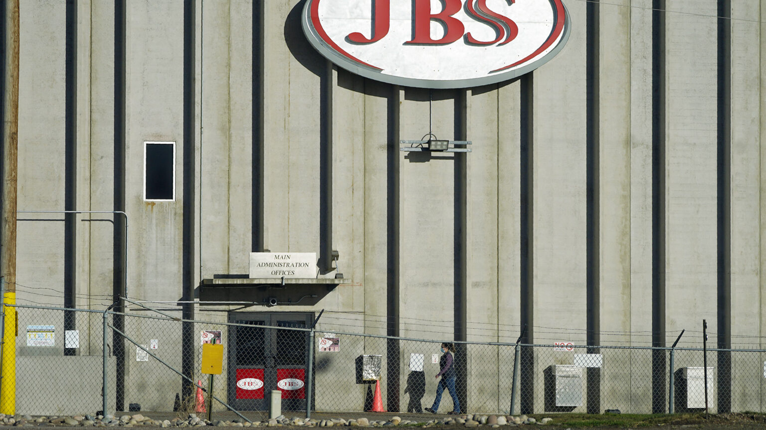 A person walks under a sign with a wordmark logo that reads JBS toward an entrance of a multi-story building with concrete and metal walls, with a chain-link fence topped by barbed wire in the foreground.