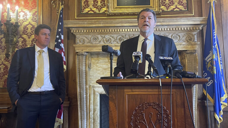Robert Wittke stands behind a wood podium and speaks into multiple microphones while Eric Wimberger stands behind and to the site, with U.S. and Wisconsin flags and a fireplace with carved stone mantel in the background.