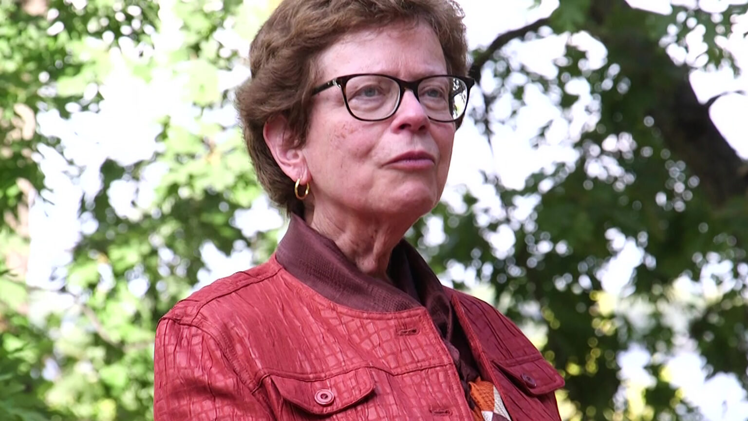 Rebecca Blank stands and speaks with out-of-focus tree foliage in the background.