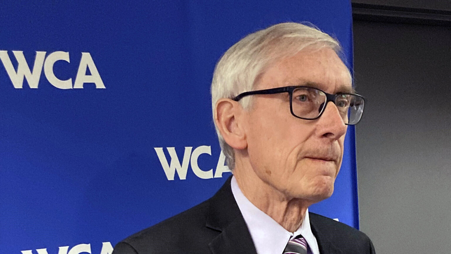 Tony Evers listens while standing in front of a logo backdrop banner with a repeated WCA wordmark.