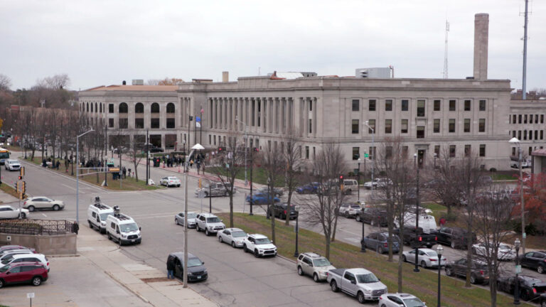 Vehicles travel and are parked along a boulevard as it intersects another road in front of a three-story masonry building with columns on the upper two stories of its façade, with groups of people standing outside its entrance and in the middle of the boulevard.