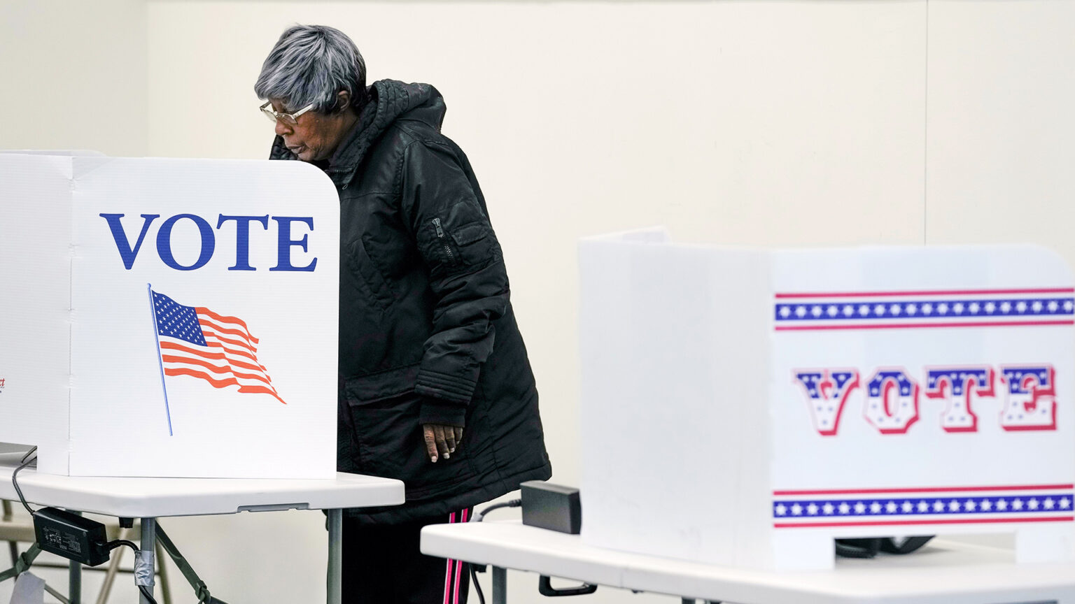 A voter stands behind a privacy screen mounted on a folding plastic table with a graphic of the U.S. flag and the word VOTE and casts a ballot, with another table with a similar screen and different graphic in the foreground.