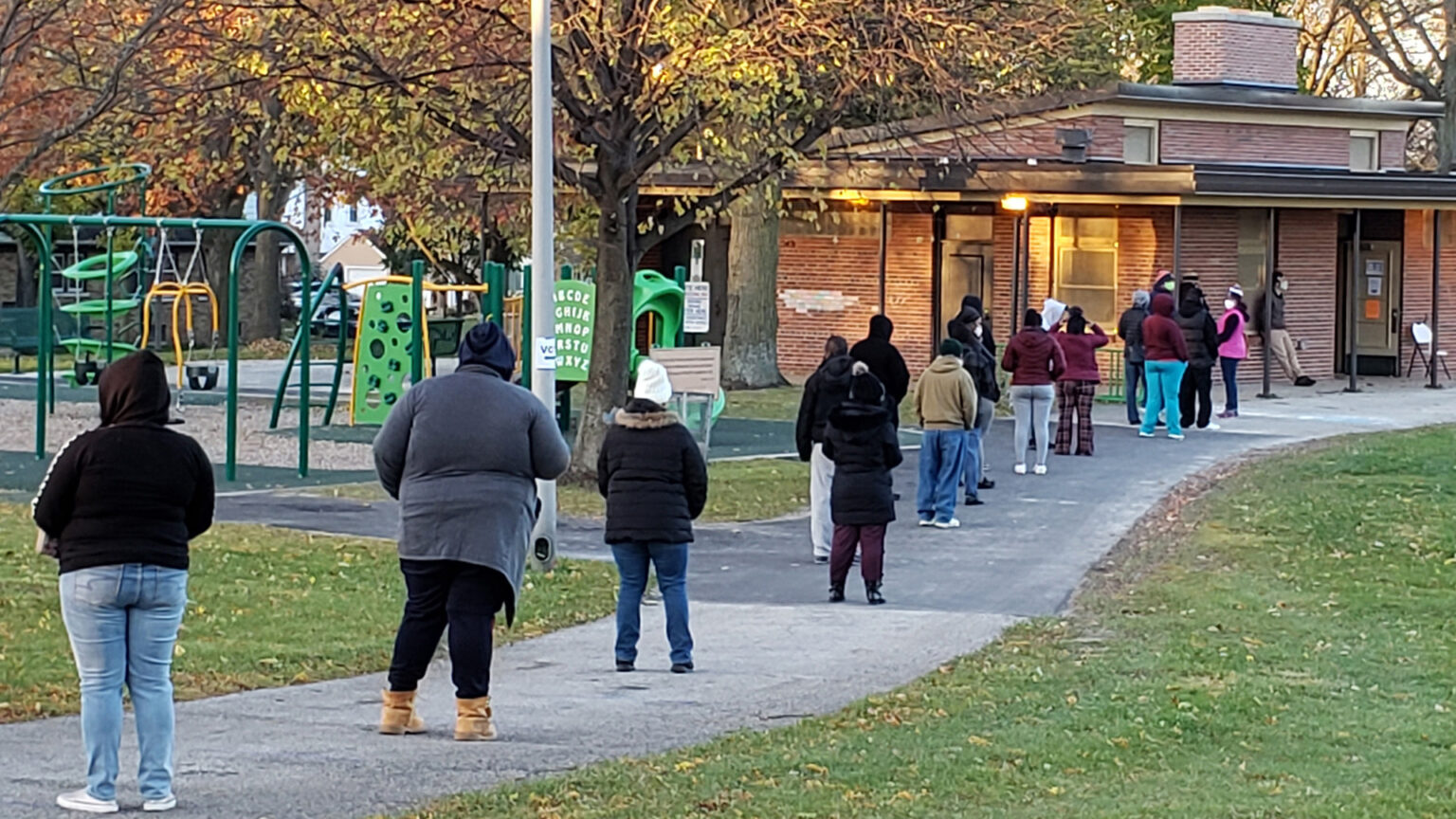 People stand in a line with wide spaces between each individual on a sidewalk leading toward a brick building, with playground equipment and trees on behind the queue.