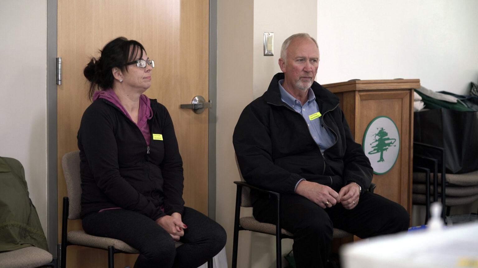 Two people sit in stackable chairs, with more chairs on either side of them and a closed door and podium behind them.