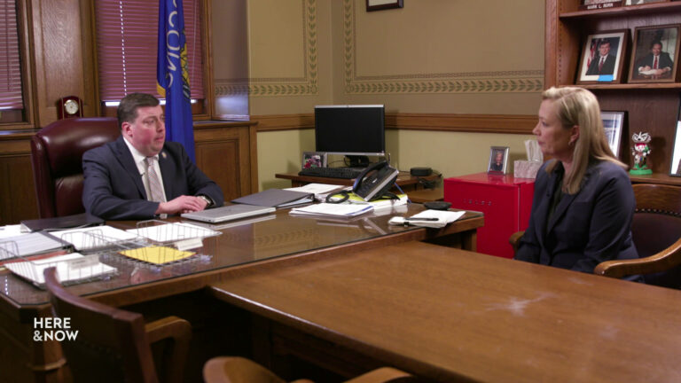 Mark Born and Frederica Freyberg sit in chairs in an office with a Wisconsin flags, a bookcase with portraits, and a computer desk with keyboard and screen in the background, and a meeting table with more chairs in the foreground.