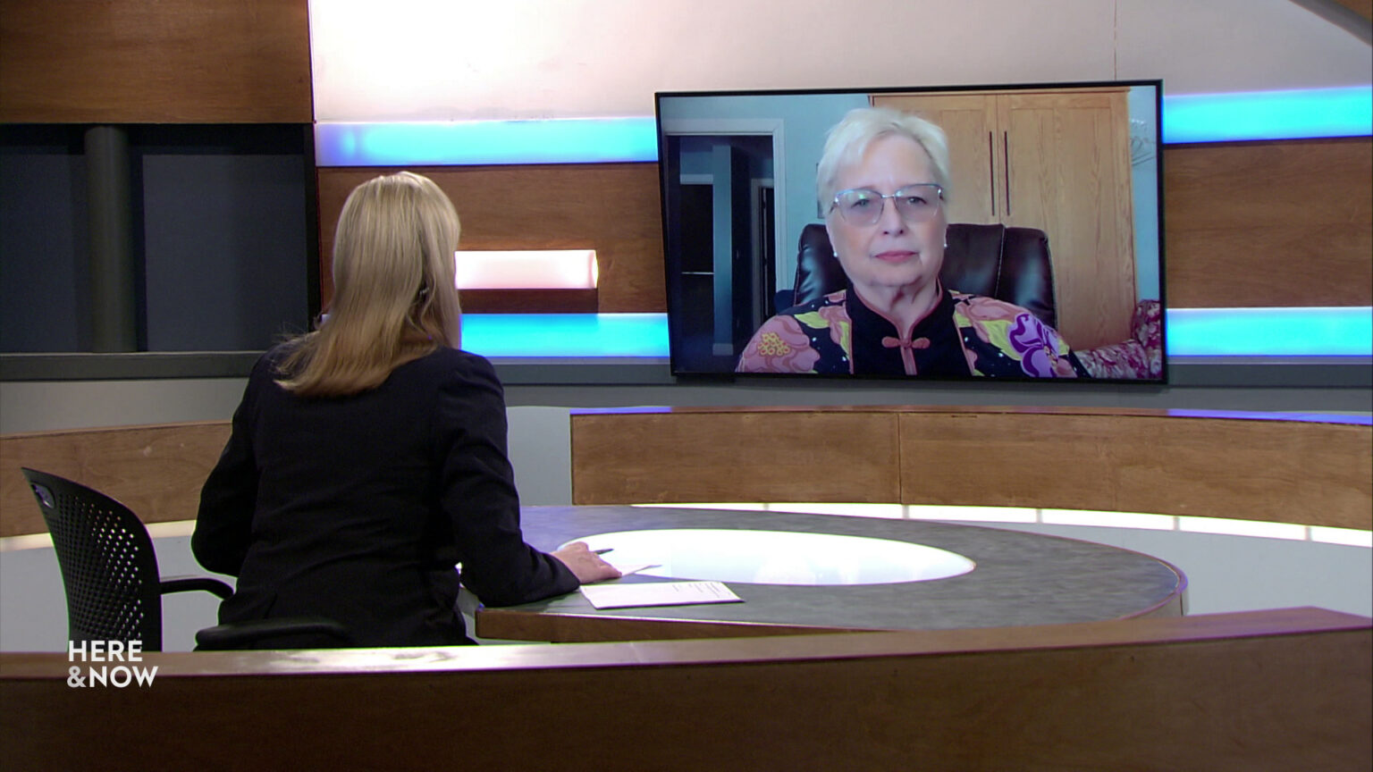 Frederica Freyberg sits at a desk on the Here & Now set and faces a video monitor showing an image of Janine Geske.