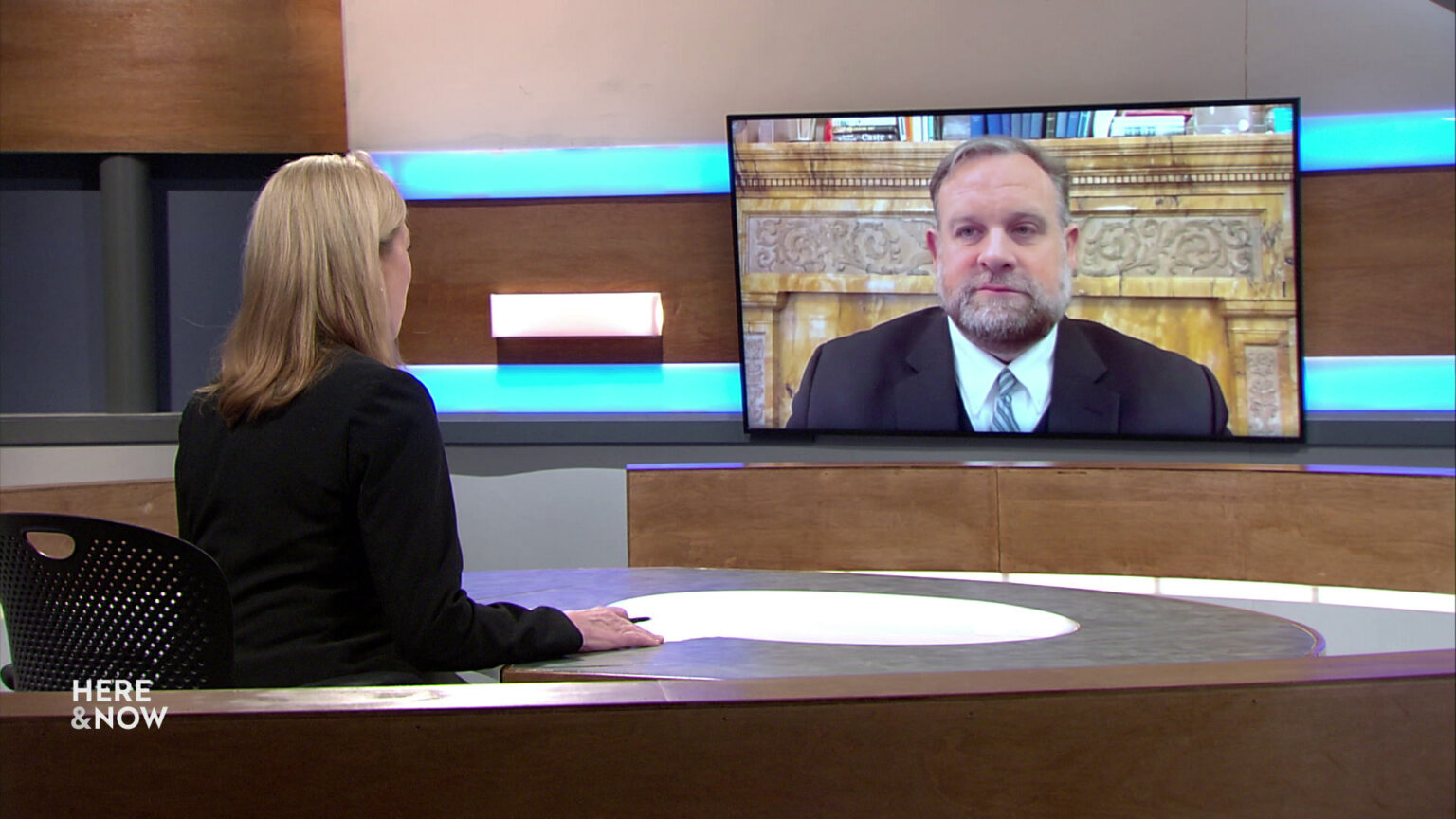 Frederica Freyberg sits at a desk on the Here & Now set and faces a video monitor showing an image of Cory Mason.