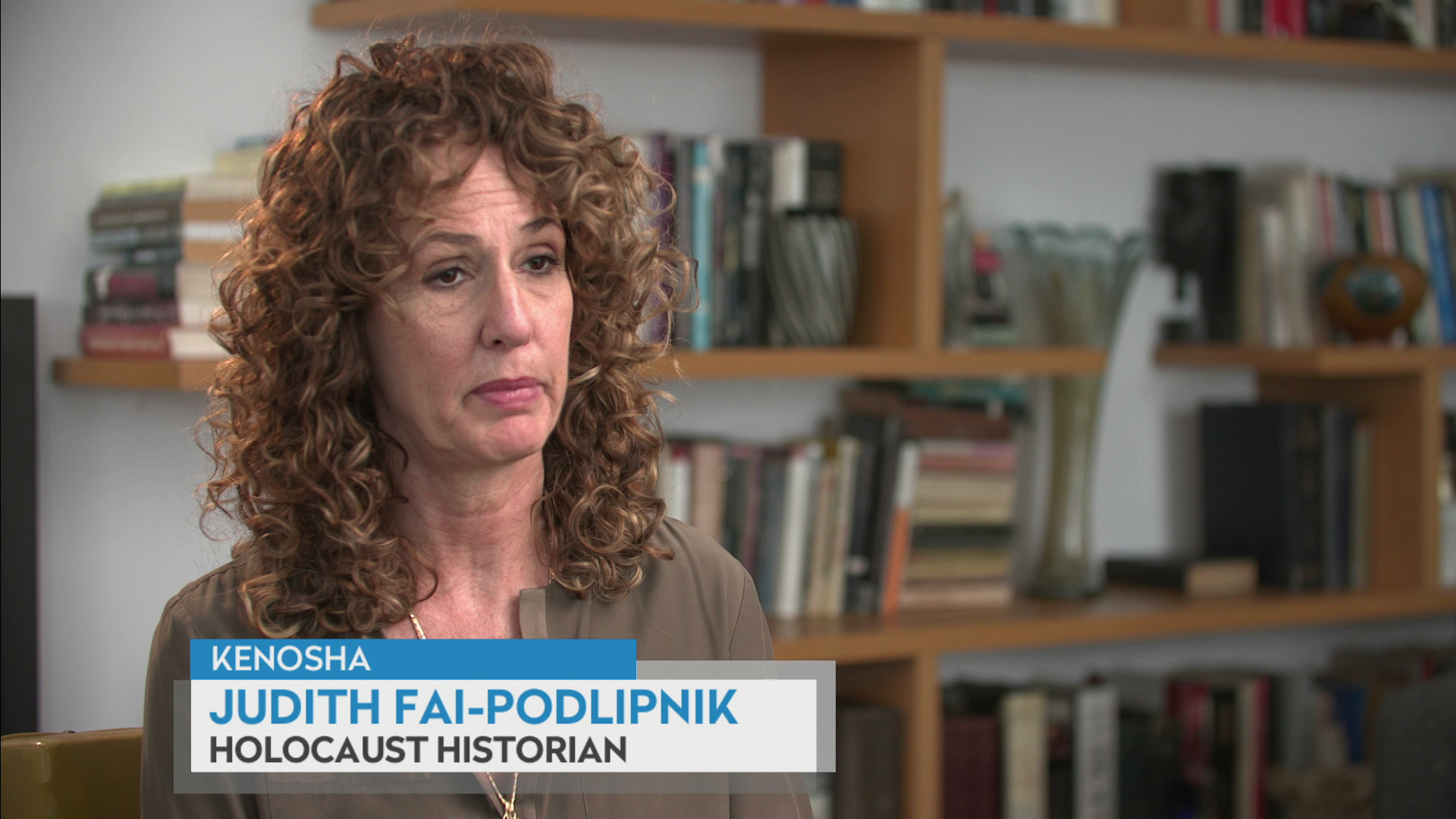 A still image from a video interview shows Judith Fai-Podlipnik seated in a room with sheving covered with books, pottery and other items in the background, with a graphic at bottom reading "Kenosha," "Judith Fai-Podlipnik" and "Holocaust Historian."