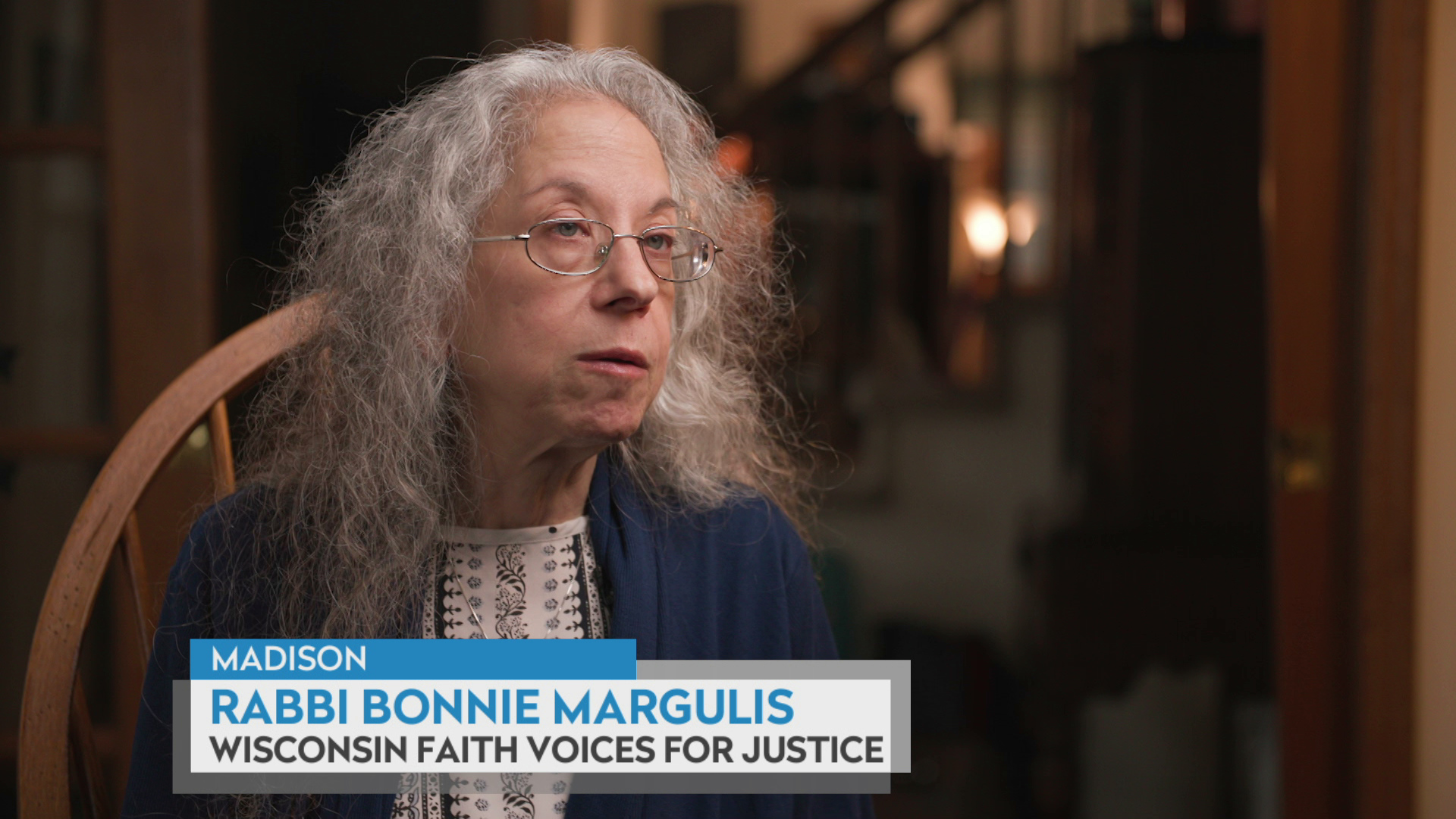 Bonnie Margulis sits on a chair with open double doors and a stairway in the background, with a graphic at bottom reading "Madison," "Rabbi Bonnie Margulis," and "Wisconsin Faith Voices for Justice."