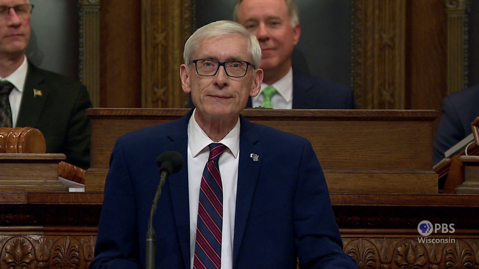 Tony Evers stands and speaks into a microphone with Robin Vos seated at a dais behind him.