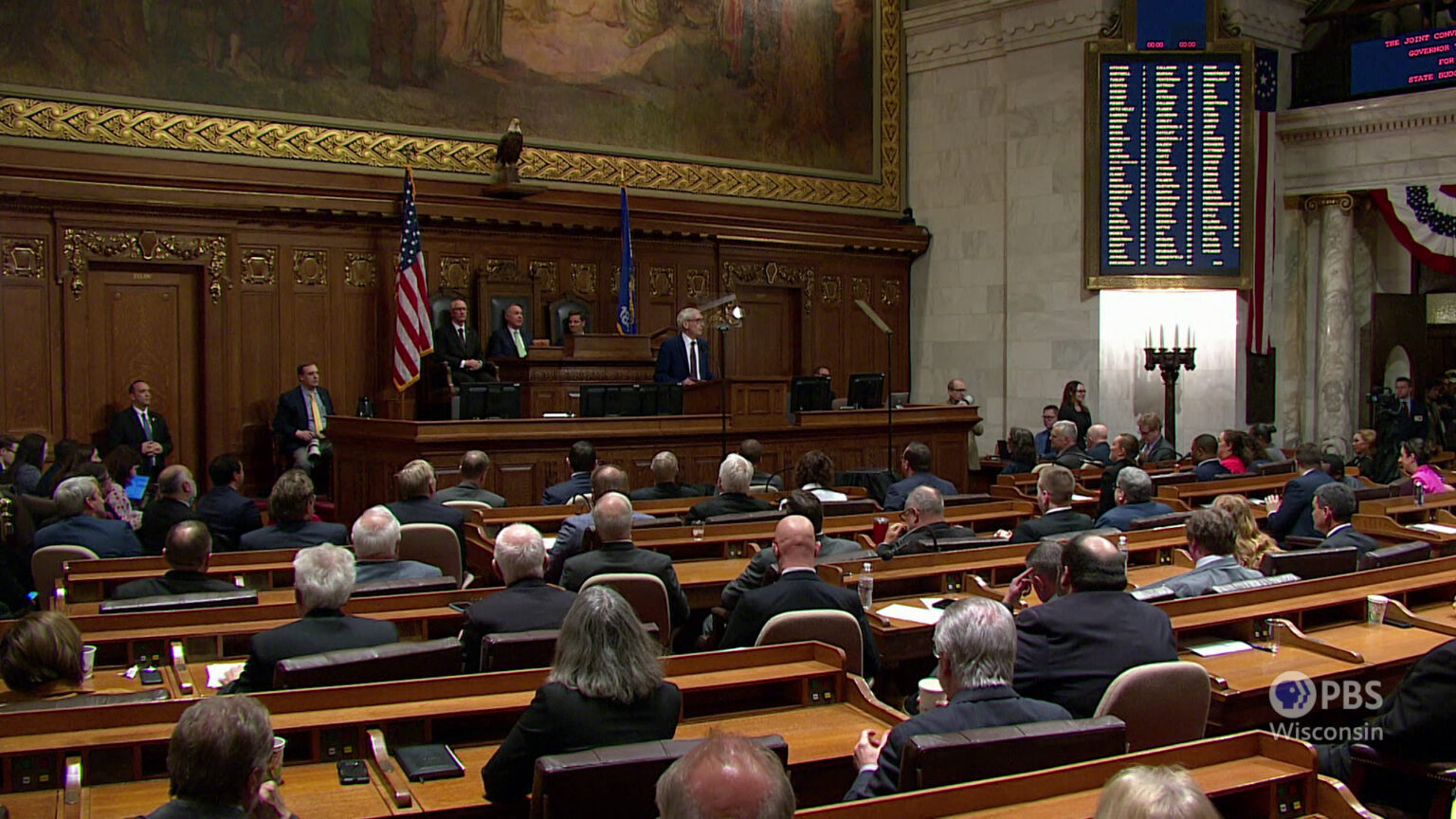 People seated in multiple rows of wooden desks face a dais where three people sit in high-backed chairs behind Tony Evers, who stands behinds a podium and speaks, with a U.S. and Wisconsin flag, carved wood panels, a taxidermy bald eagle and a large painting on the wall behind them, with an electronic vote display mounted on an adjacent marble block wall, and more people seated and standing on either side.