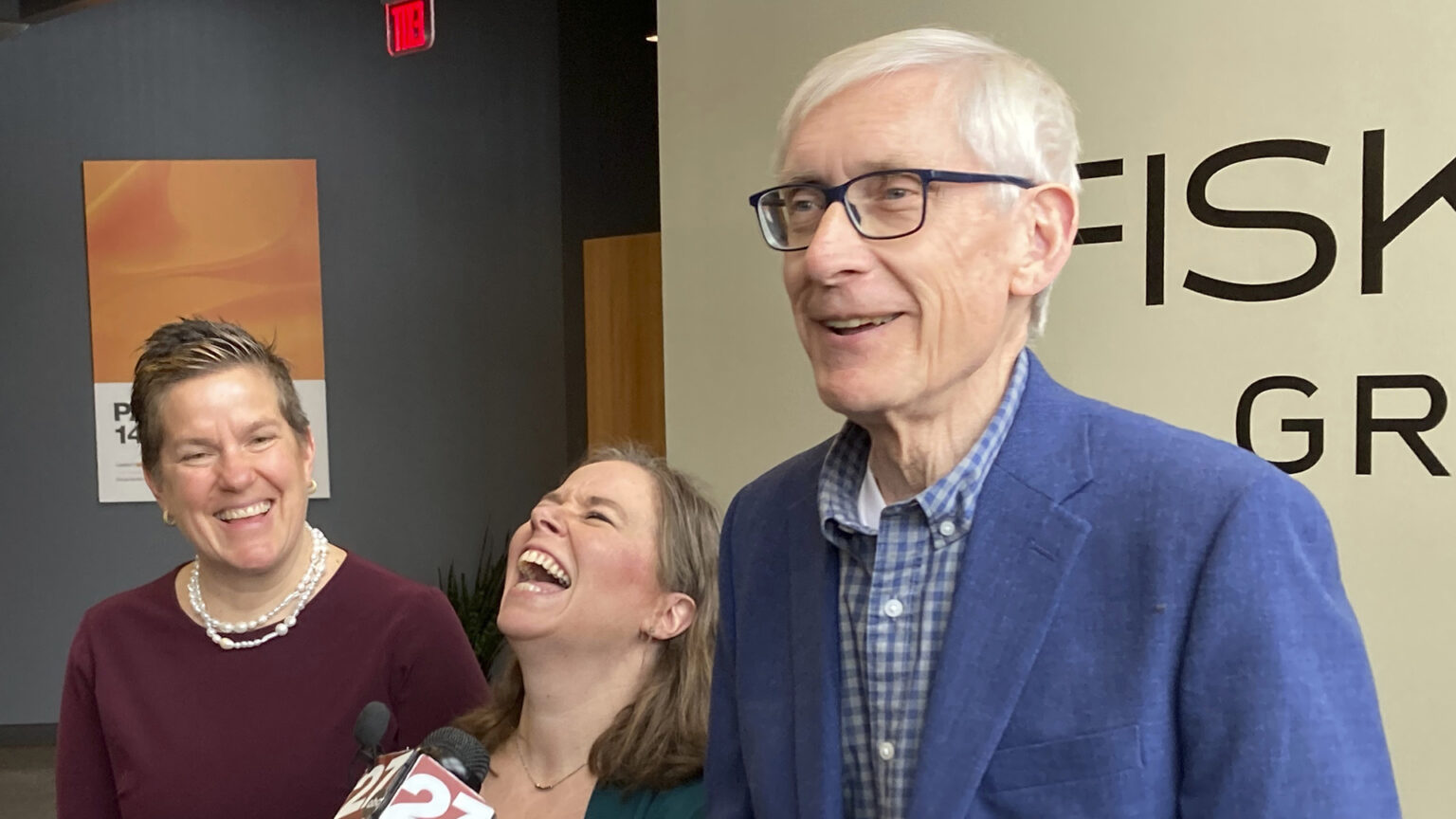 Missy Hughes and Tony Evers stand on other side of Sara Rodriguez, who is laughing while speaking into a microphone, in a room with two walls painted different colors.