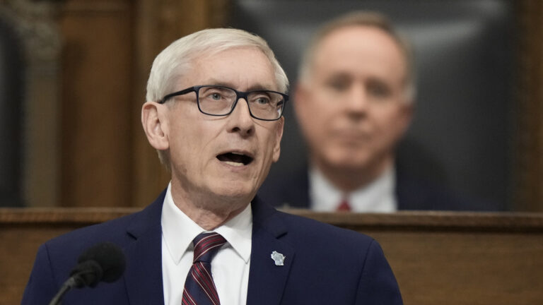 Tony Evers stands and speaks behind a microphone with Robin Vos, appearing out-of-focus, seated behind on a dais.