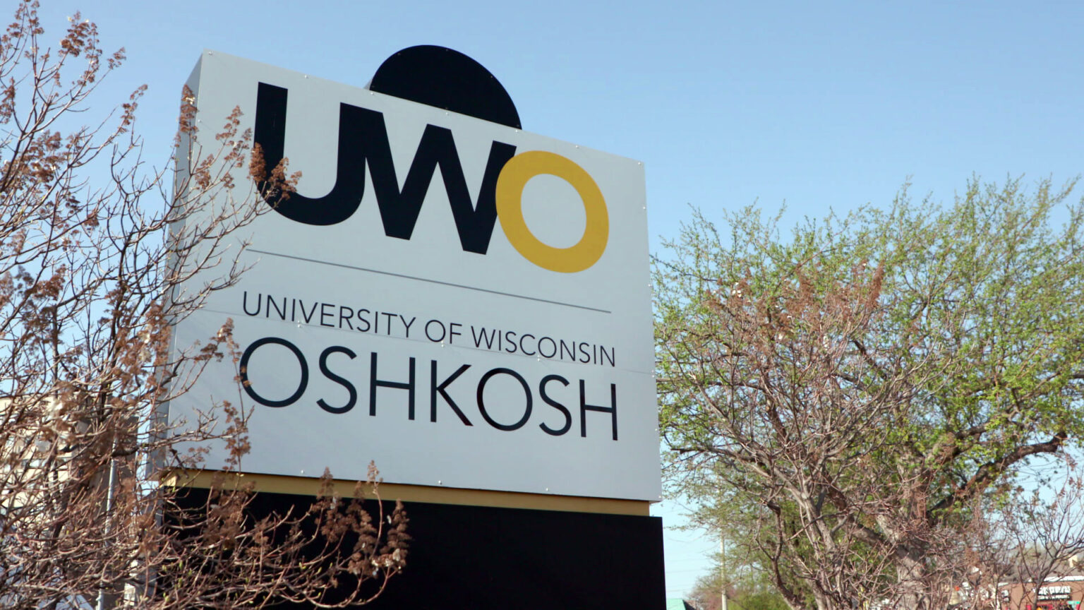 A sign with a stylized wordmark incorporating the letters UWO and the words University of Wisconsin and Oshkosh stands amid trees showing new spring growth in their foliage.