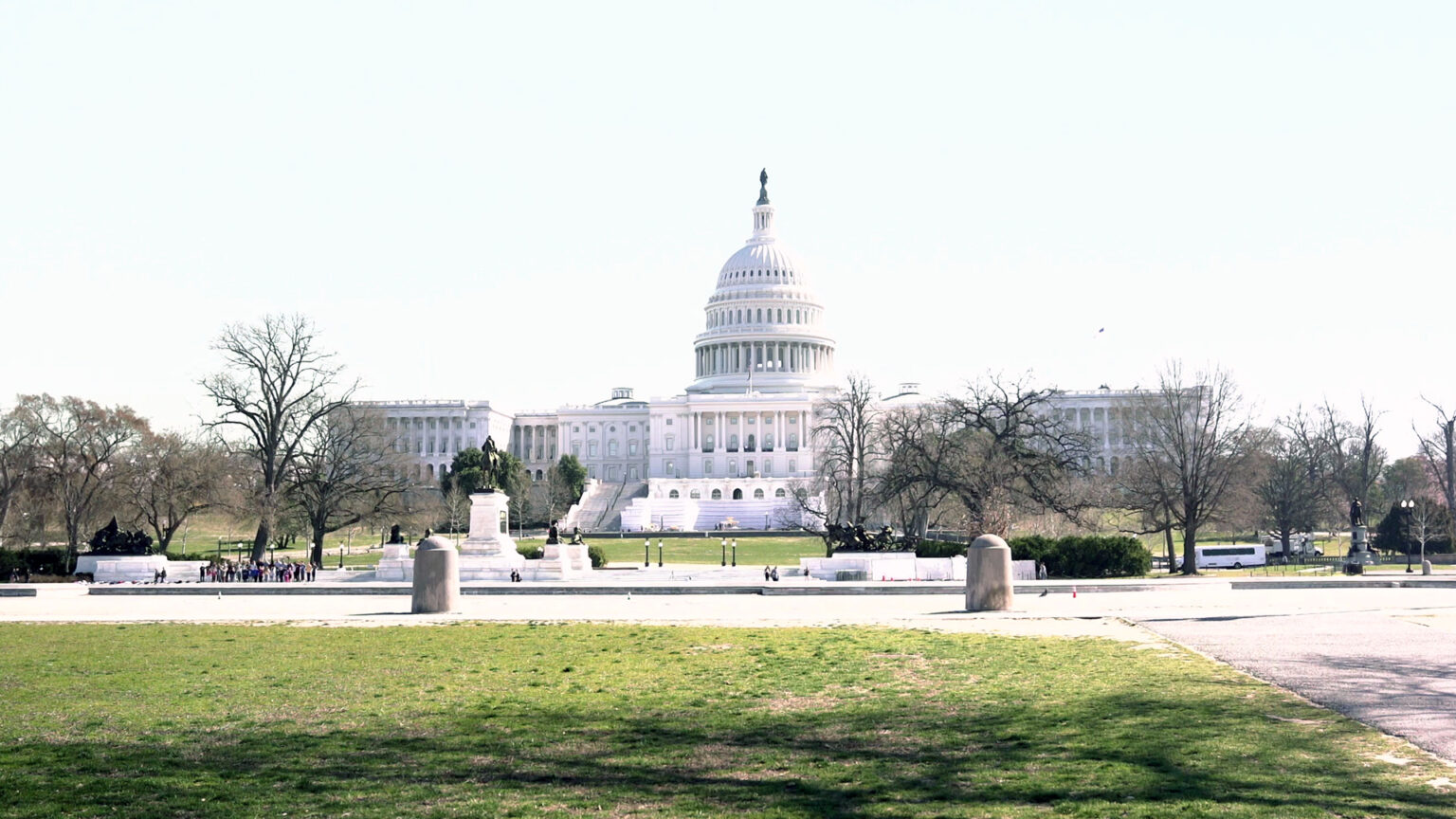 The U.S. Capitol Building stands under a cloudless ski with roads, lawns, statuary, leafless trees and pedestrians in the foreground.