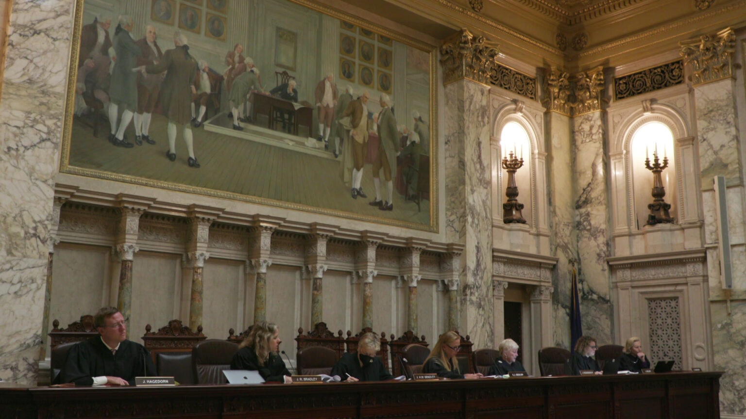 The seven justices of the Wisconsin Supreme Court sit on high-backed wood-and-leather chairs behind a court dais in a room with marble walls, crown moulding, light fixtures set within niches and a large painting.