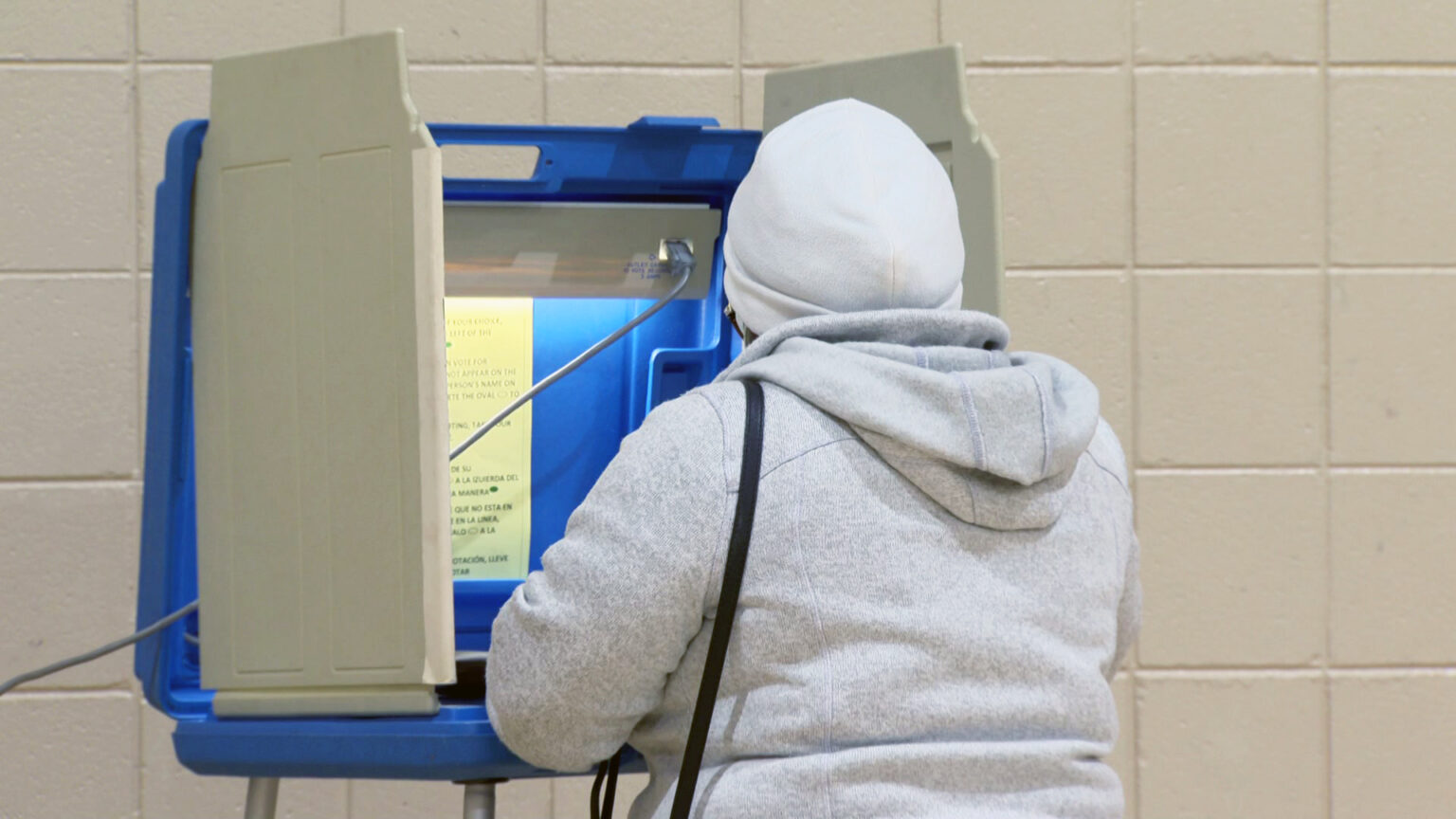 A voter fills out their ballot while standing at a voting booth with plastic privacy dividers, a light and vote procedure instructions, with a painted concrete block wall in the background.