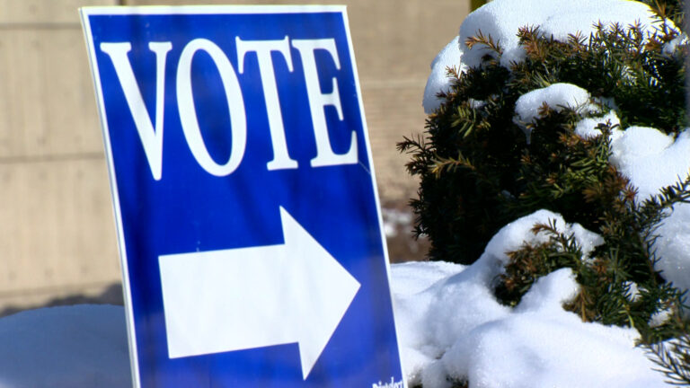 A yard sign with the word Vote and an arrow stands next to a snow-covered shrub, with concrete and brick walls in the background.
