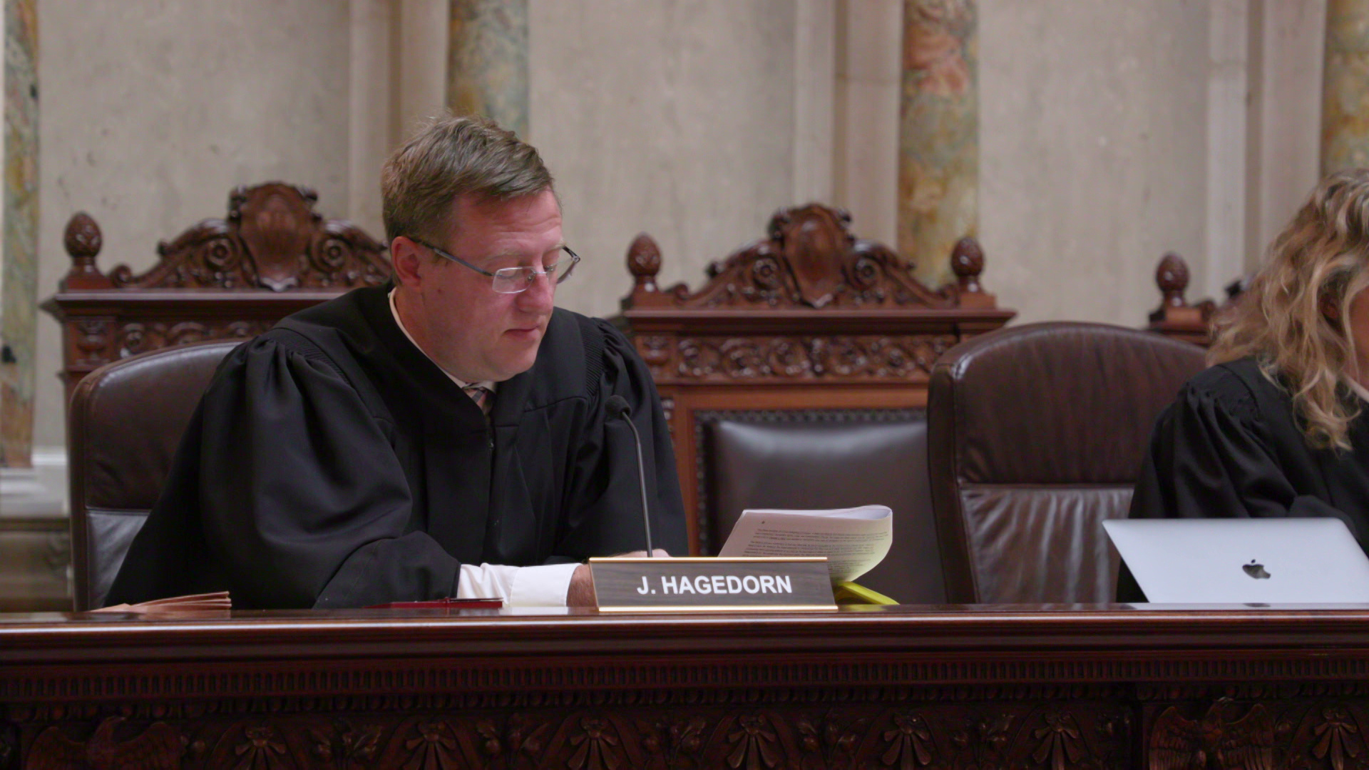 Brian Hagedorn, wearing judicial robes, looks at a paper document and speaks while sitting in a high-backed leather and wood chair behind a dais with a nameplate reading "J. Hagedorn" in a room with marble walls and pillars.