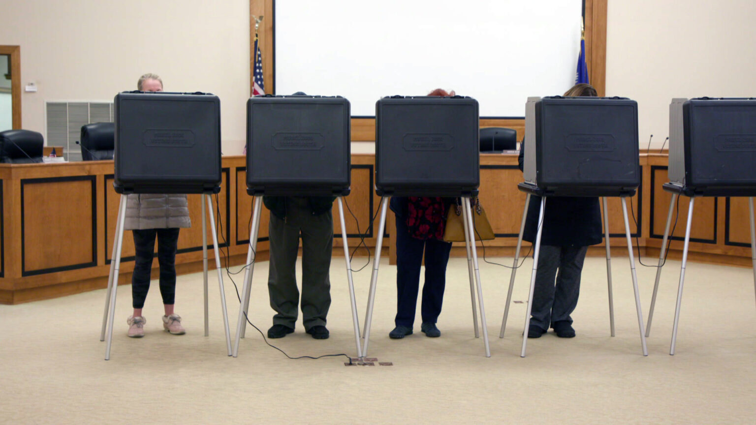 A row of collapsible voting booths with metal legs and plastic bodies stand in a row, with voters standing in four of them with only their legs visible, in a government meeting room with a chairs placed behind a low dais, the U.S. and Wisconsin flags, and a projector screen in the background.