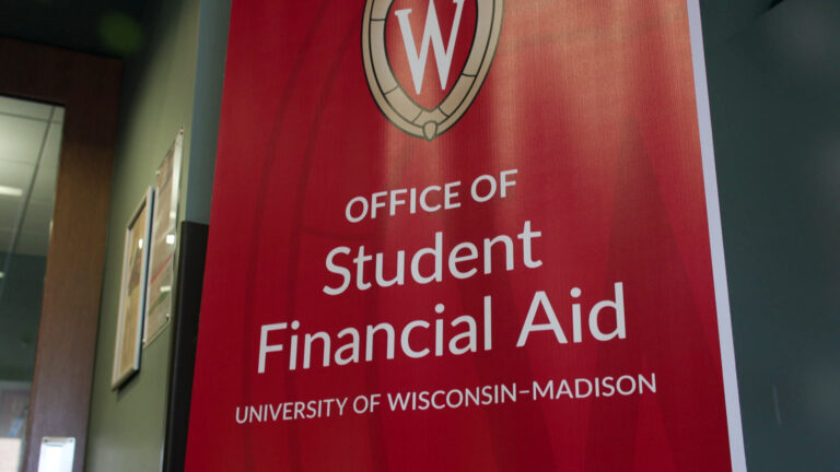 A vinyl banner with words Office of Student Financial  Aid and University of Wisconsin-Madison with the university's 'W' crest on it stands in a hallway, with a glass-paneled door in the background.