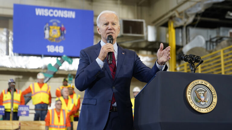 Joe Biden stands behind and to the side of a podium affixed with the presidential seal and two mounted microphones, holding a wireless microphone in his right hand and gesturing with this left hand, in a multi-story room with concrete walls, safety railings and HVAC equipment on the ceiling, with other people wearing safety vests and hard hats standing in risers in the background under a Wisconsin flag.