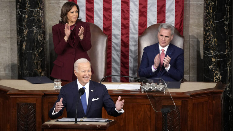 Joe Biden stands behind a podium and smiles while gesturing with both hands, while on a dais behind him Kamala Harris stands while applauding and Kevin McCarthy sits while applauding.