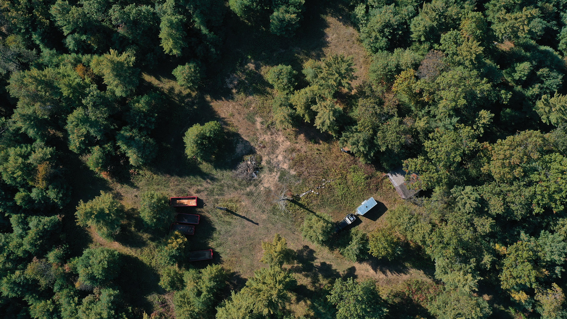 An aerial photo shows a clearing in a wooded area with multiple construction dumpsters and vehicles, with exposed soil at its center.