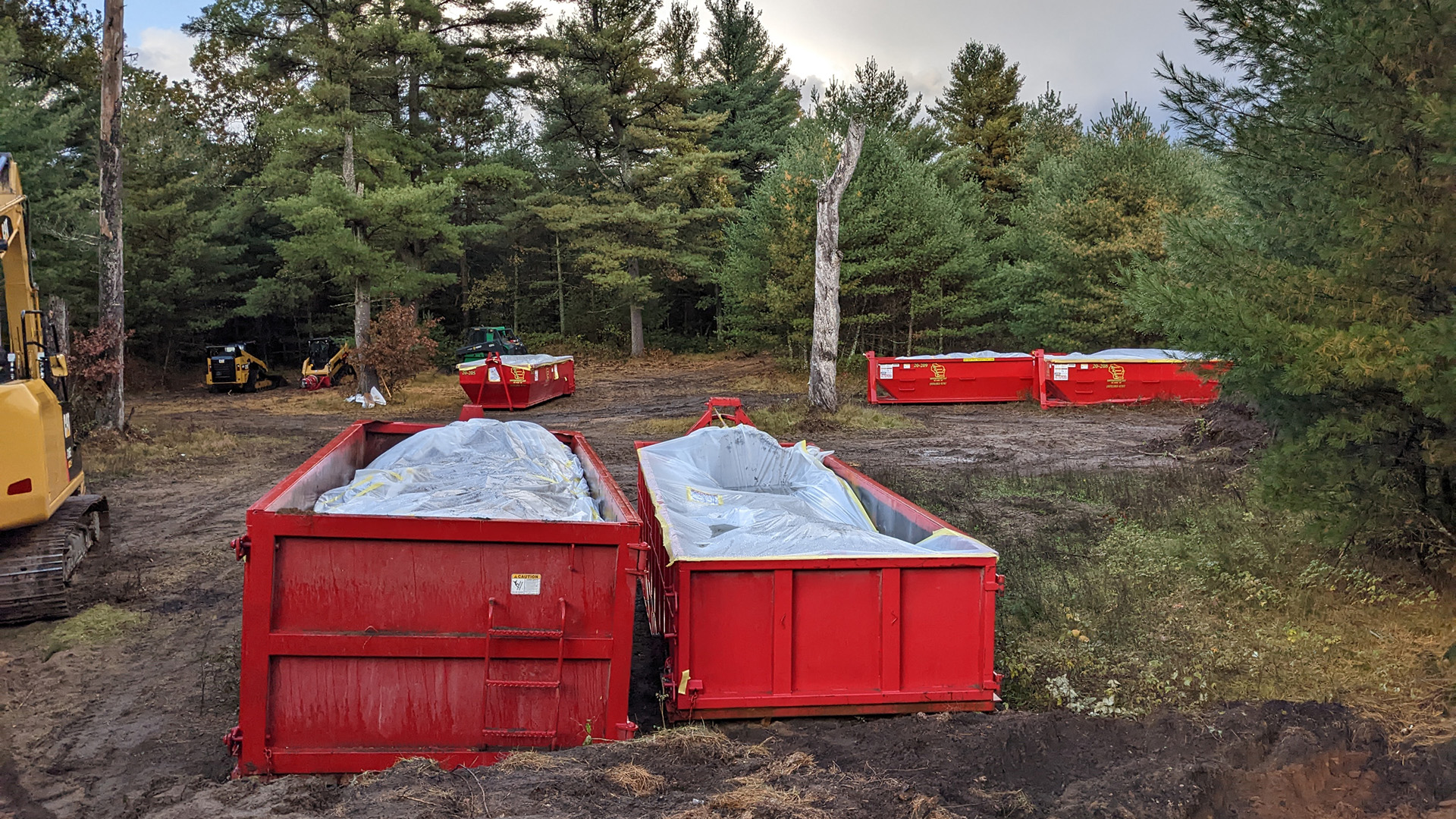 Five construction dumpsters are placed in three locations alongside multiple construction vehicles in a clearing, filled with debris covered by plastic sheets, with trees in the background.