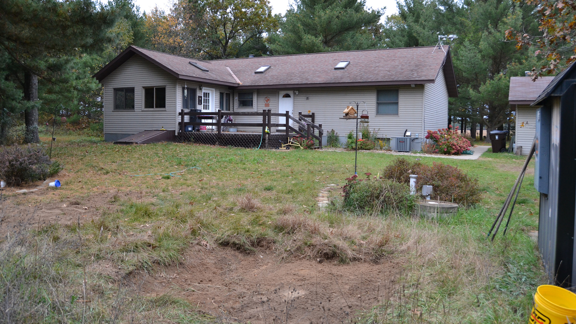 A single-story house with a deck, skylights and exterior basement entrance stands in a clearing alongside multiple outbuildings and in front of a line of trees.