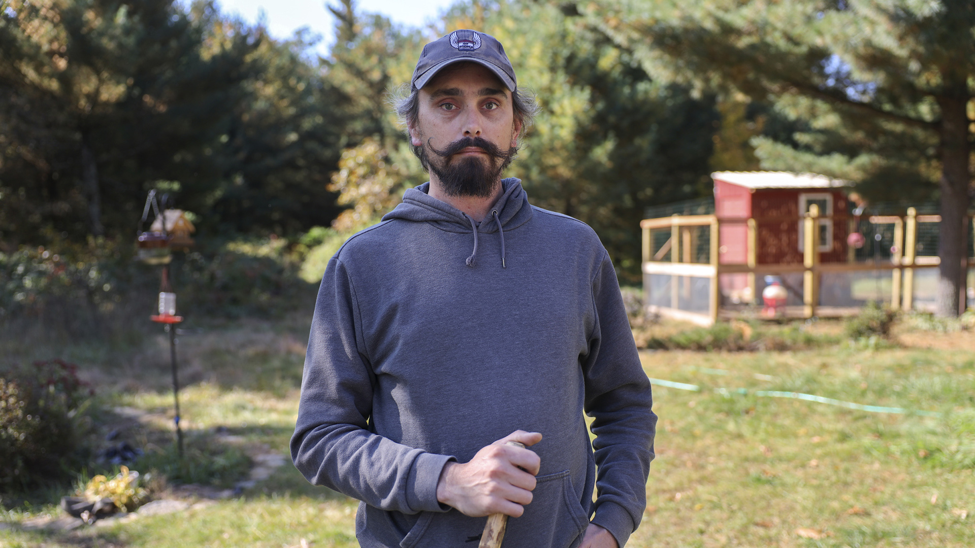 Zach Skrede stands in a yard with a bird feeder, small animal enclosure, hose and trees in the background.