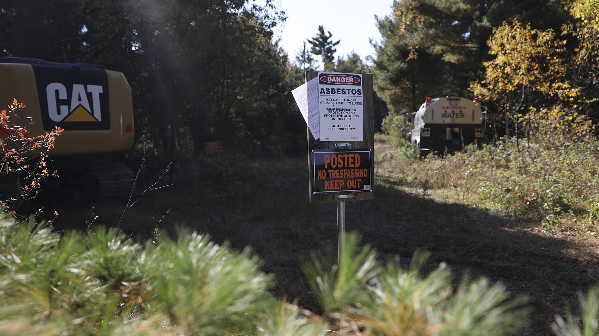 Two warning signs are mounted on a board placed next to a dirt track, with one featuring a "Danger" symbol regarding asbestos and another reading "Posted" and "No Trespassing Keep Out," with a water tank truck, construction vehicle and trees in the background.