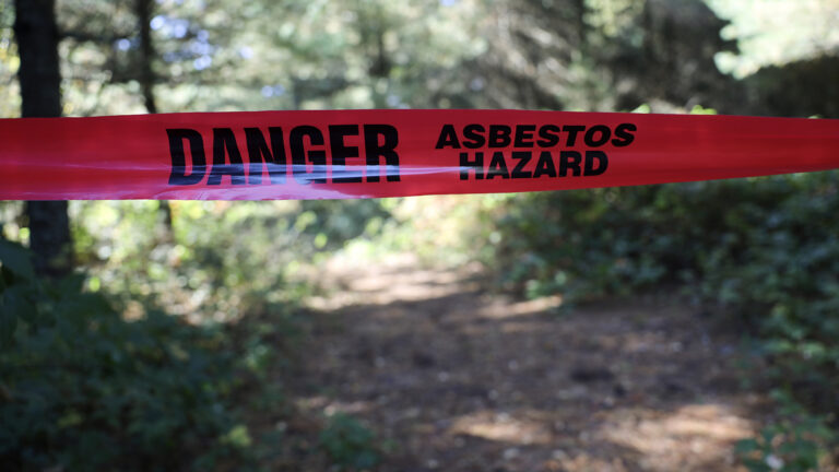 Plastic tape bearing the words Danger and Asbestos Hazard crosses a path in a wooded area, with sunlight trees in the background.