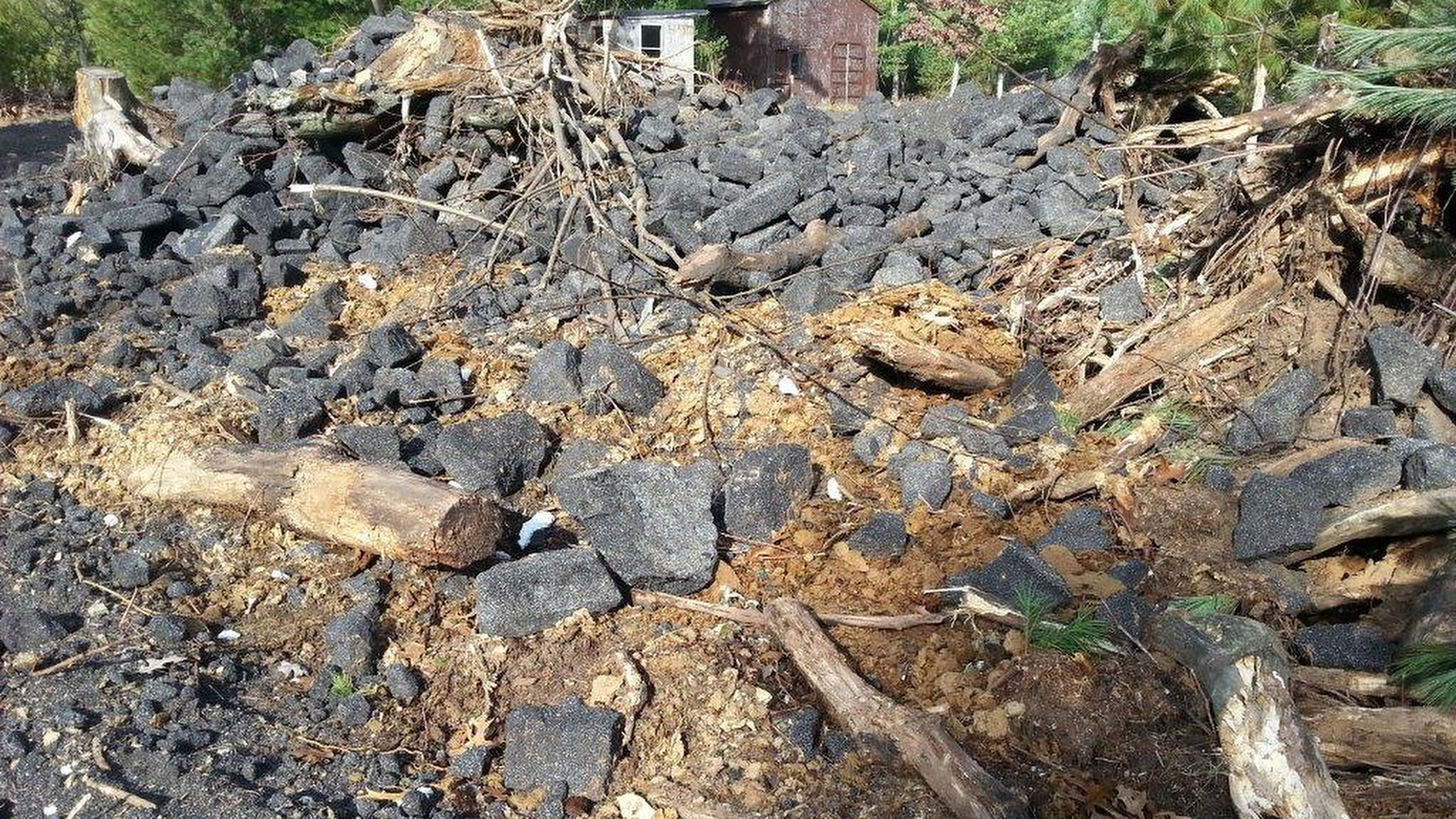 A pile of debris consisting timber fragments, sawdust and broken pieces of granular construction material sits in a pile next to trees, with buildings and more trees in the background.