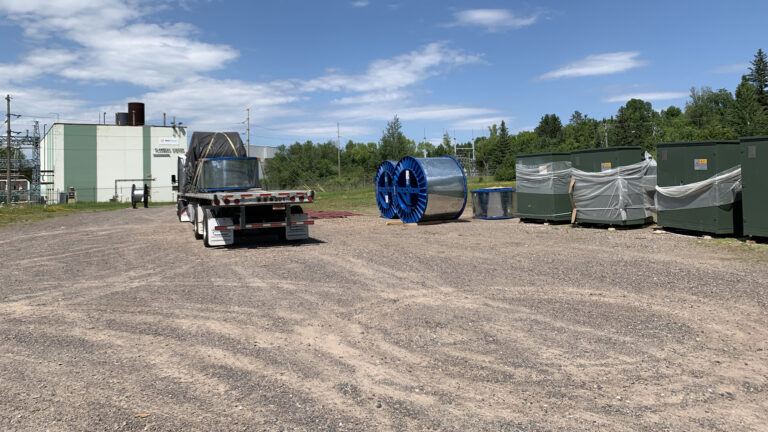 A semi truck with a spool of material strapped on its flatbed trailer is parked in a gravel lot with two other spools and multiple closed metal crates partially wrapped in a filmy material, with trees and a metal-sided industrial building with exterior electrical infrastructure in the background.