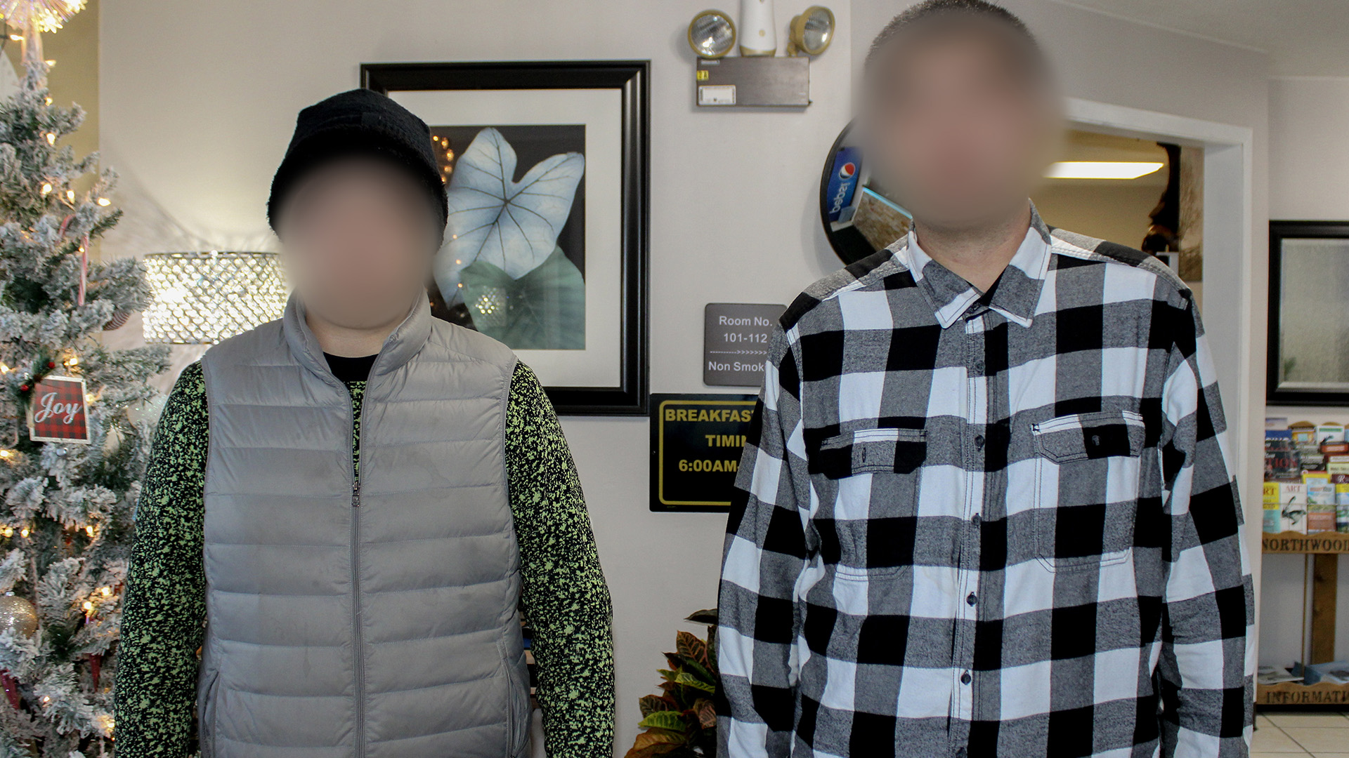 Aaron and Justin, appearing with blurry spots over their faces, posed for a portrait in a room with a Christmas tree, rack of tourism pamphlets and signs referencing motel room locations and breakfast times.