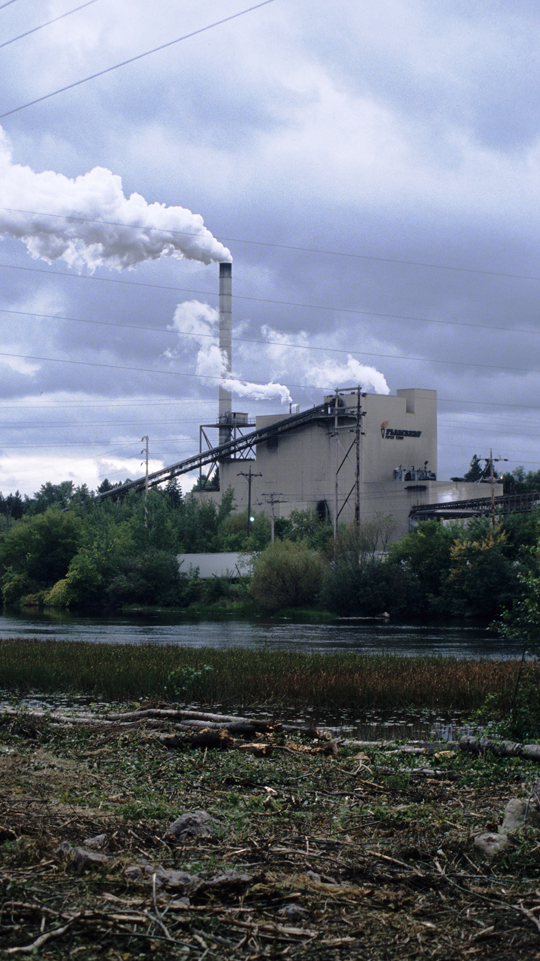 A paper mill with a tall stack and multi-story conveyer system stands amid trees on the far side of a river, with power lines crossing overhead in the foreground.