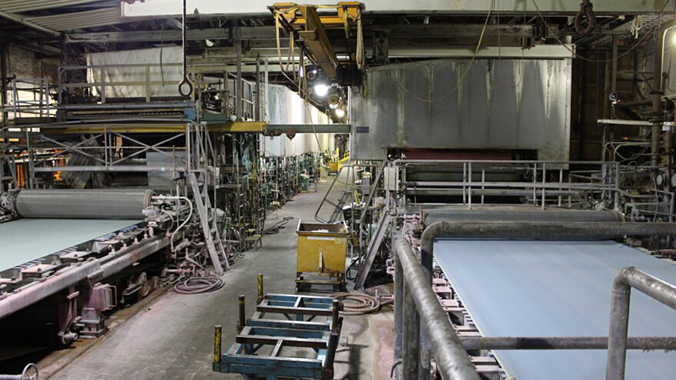 Industrial manufacturing equipment, including conveyer belt and roller components, is arrayed in two lines on the floor of a paper mill.