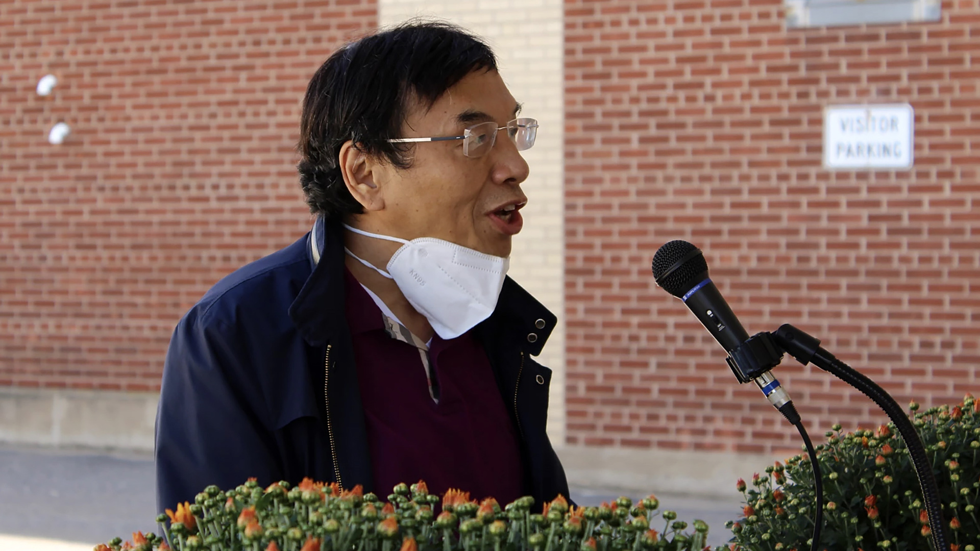 Yong Liu sits outdoors behind two bunches of flowers and speaks into a microphone, with a brick wall and a sign reading "Visitor Parking" in the background.
