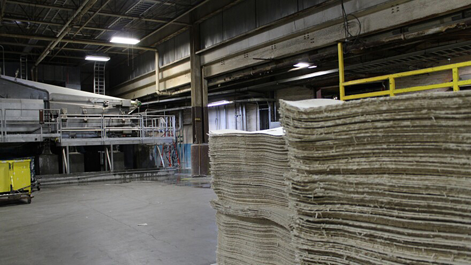 Two stacks of a paper manufacturing product stand on the concrete floor of a mill, with industrial equipment and building infrastructure in the background