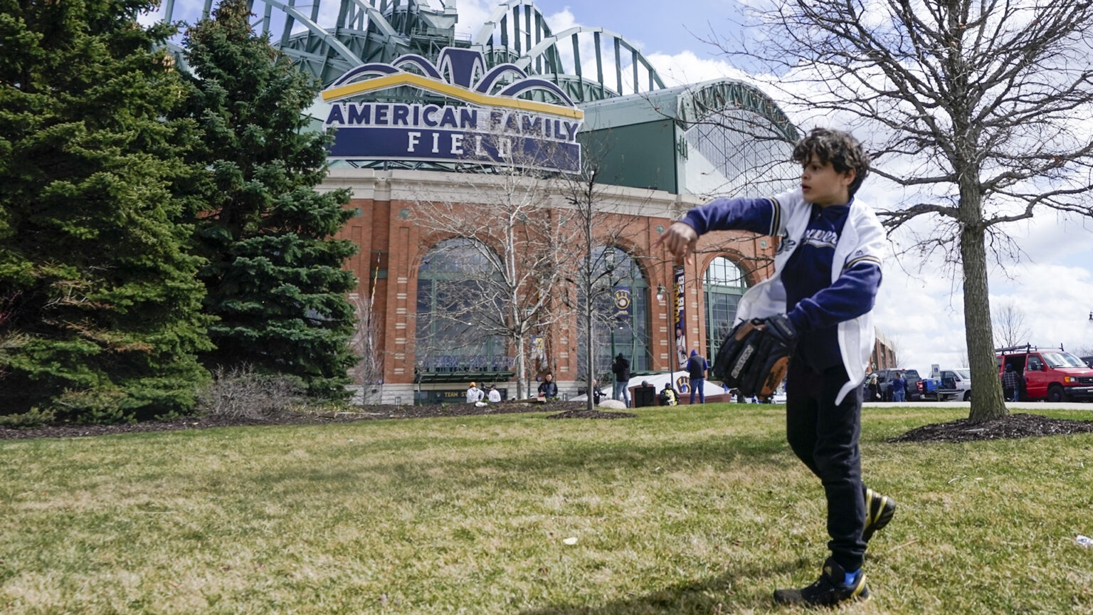 Lennon Jones throws with his right arm while standing on a lawn with several trees and the American Family Field sign on the Milwaukee Brewers stadium in the background.