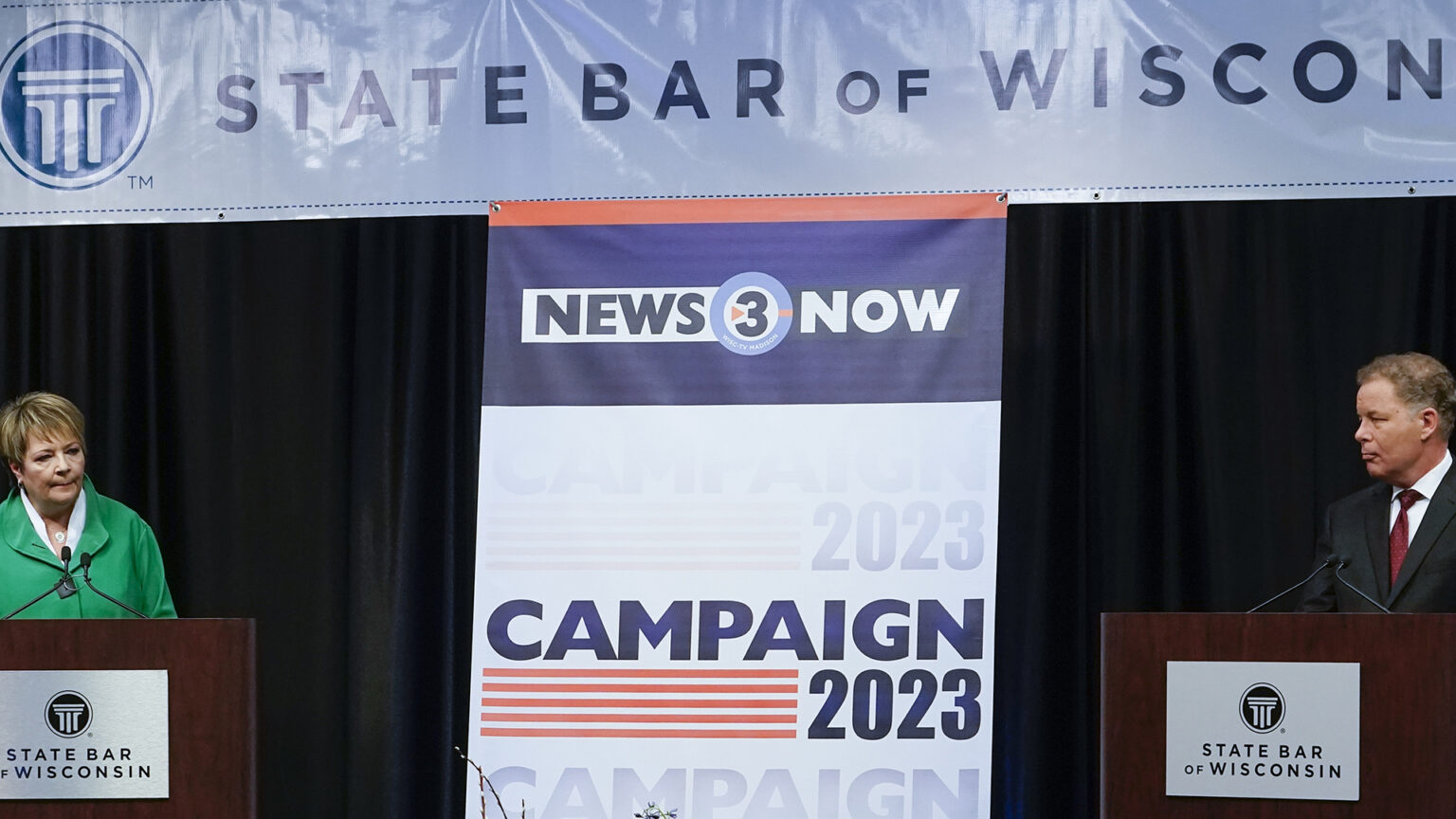 Janet Protasiewicz and Daniel Kelly stand behind podiums with microphones and labels reading State Bar of Wisconsin, with a stage curtain, horizontal banner reading State Bar of Wisconsin and vertical banner reading News 3 Now and Campaign 2023 in the background.
