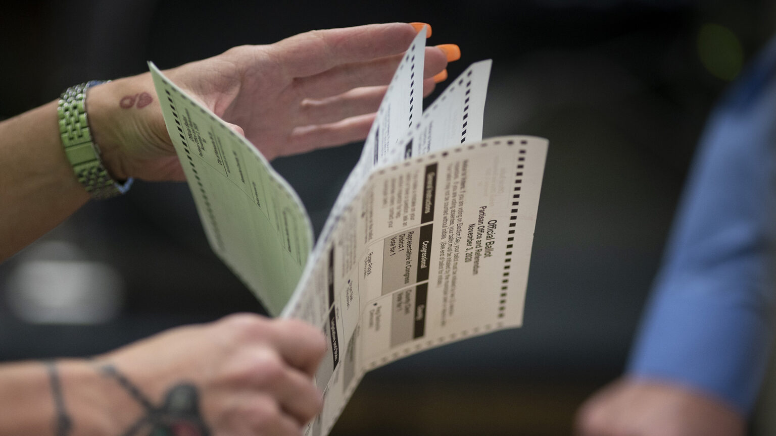 A person holds several ballots in their right hand while gesturing with their left hand.