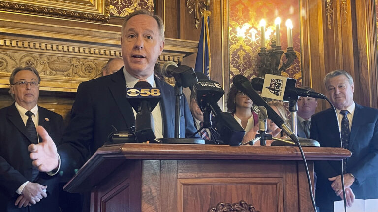 Robin Vos stands behind a podium with multiple microphones mounted on it and gestures with his right hand, with other people standing in the background in a room with toile wallpaper, wood moulding and an electric wall sconce.